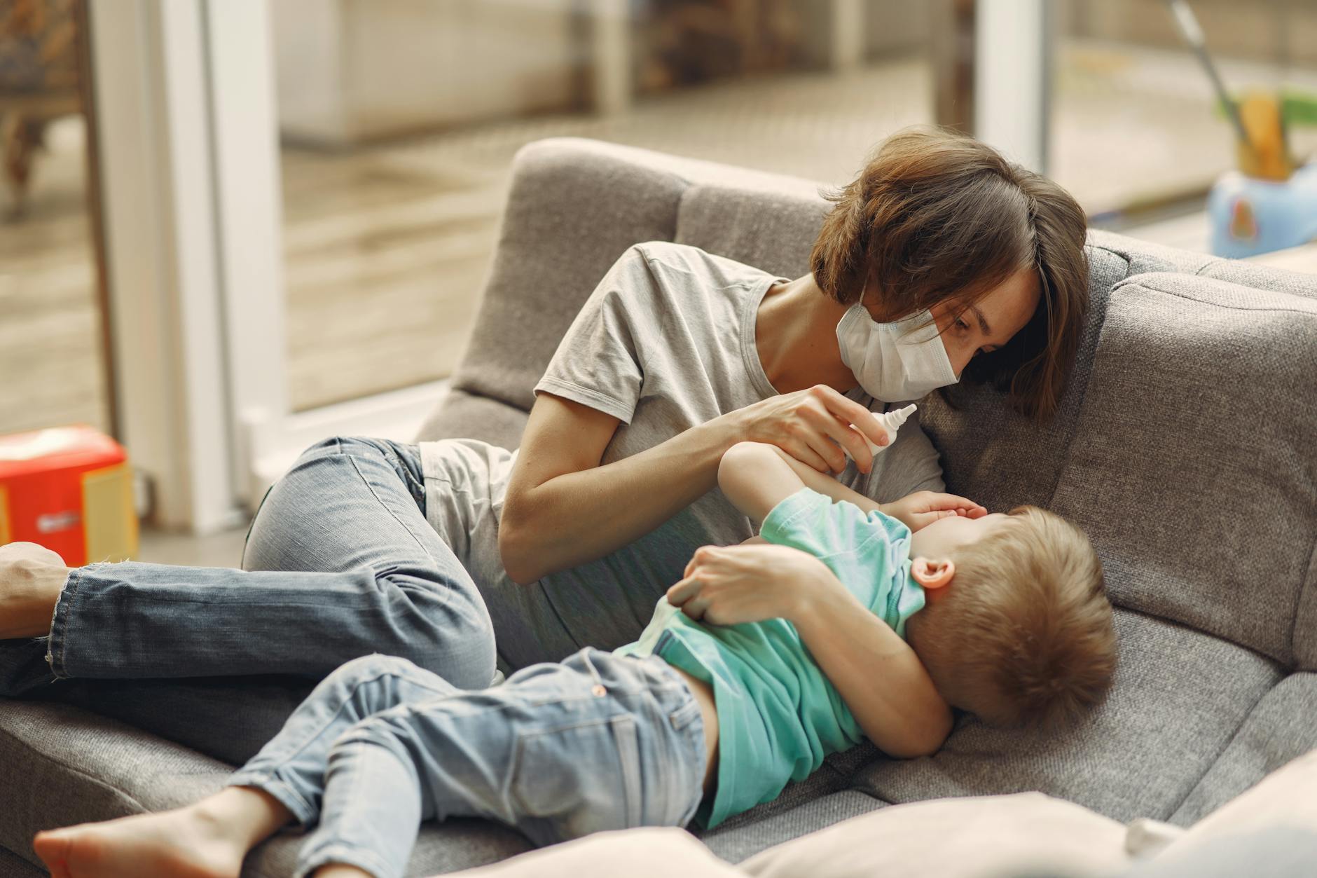 Mama w maseczce zakrapla krople do nosa choremu dziecku na kanapie