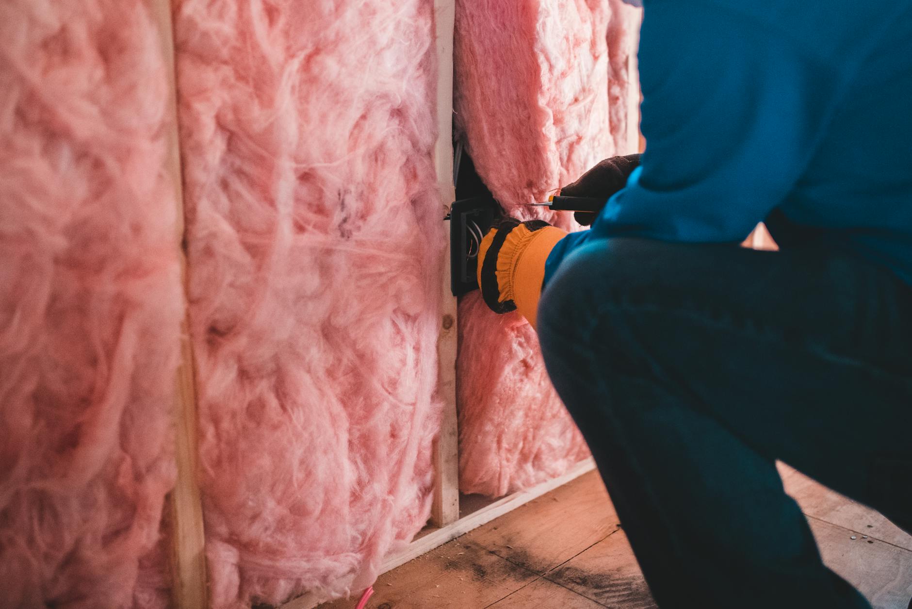 Ocieplenie domu styropianem czy wełną mineralną – jakie rozwiązanie wybrać? Pracownik w rękawicach montuje różową wełnę mineralną w ścianie