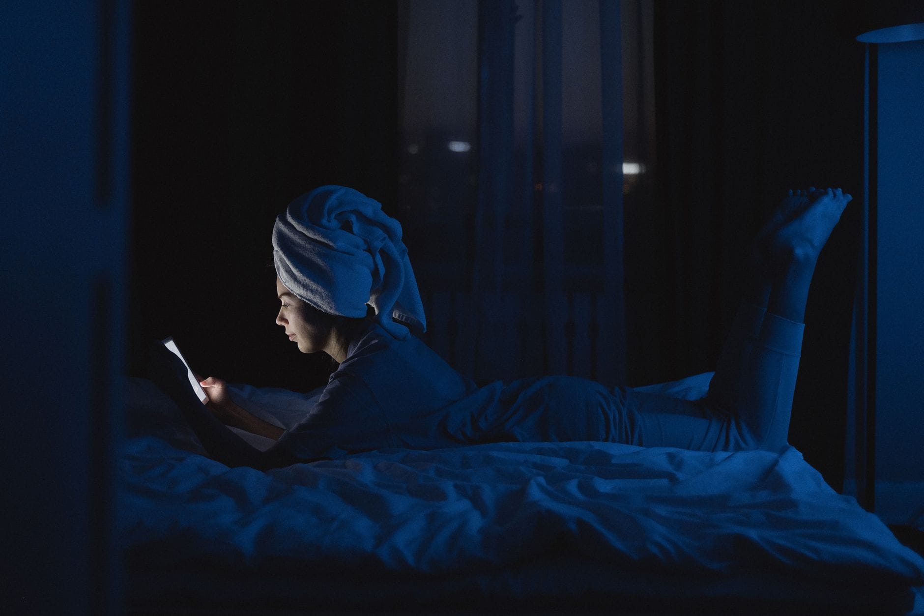 Kobieta leżąca nocą w łóżku z ręcznikiem na głowie i telefonem w dłoni